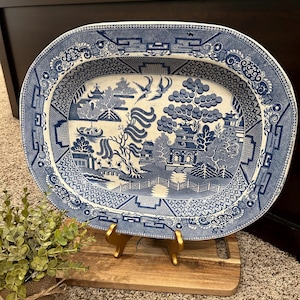 May include: A blue and white oval serving platter with a traditional Chinese willow pattern. The platter is on a wooden stand with gold metal legs.
