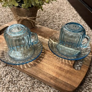 May include: Two light blue glass teacups with matching saucers on a wooden tray. The cups have a ribbed design and are sitting on a wooden surface.