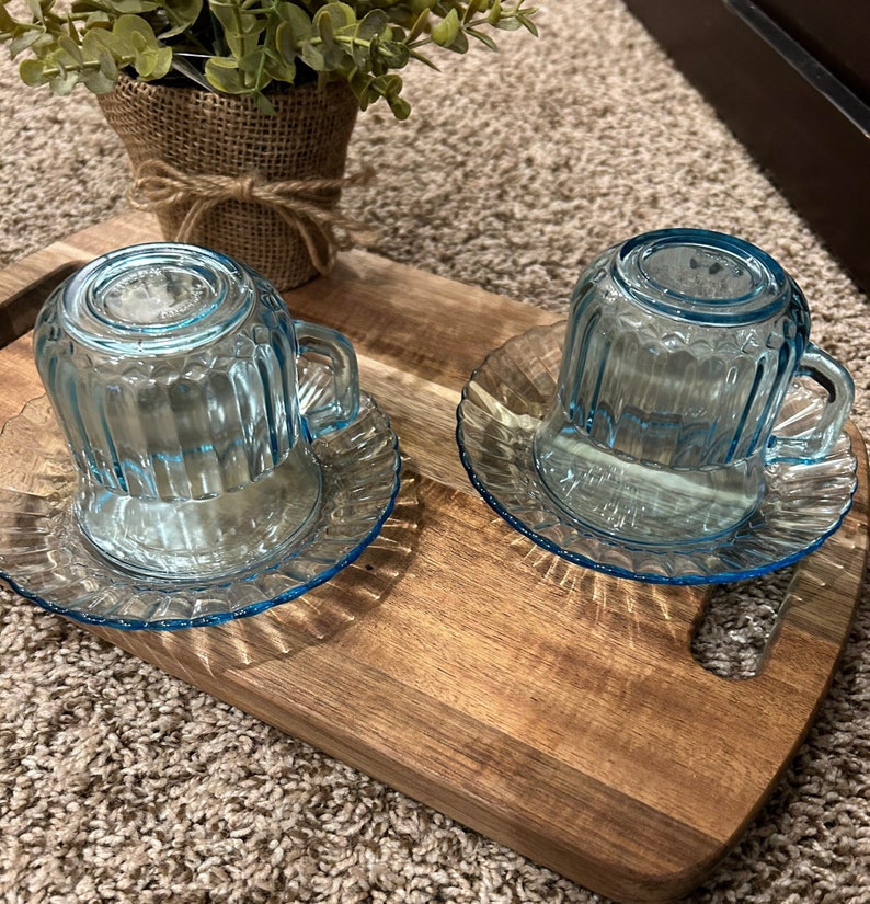 May include: Two light blue glass teacups with matching saucers on a wooden tray. The cups have a ribbed design and are sitting on a wooden surface.