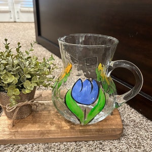 May include: A clear glass pitcher with a blue flower and yellow and green leaves painted on the side.