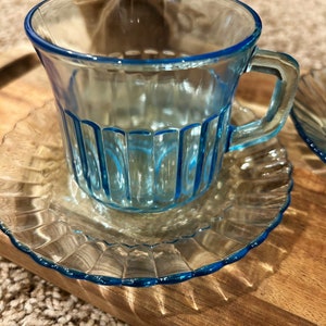May include: A light blue glass teacup and saucer set. The cup has vertical lines and a handle. The saucer has a scalloped edge.