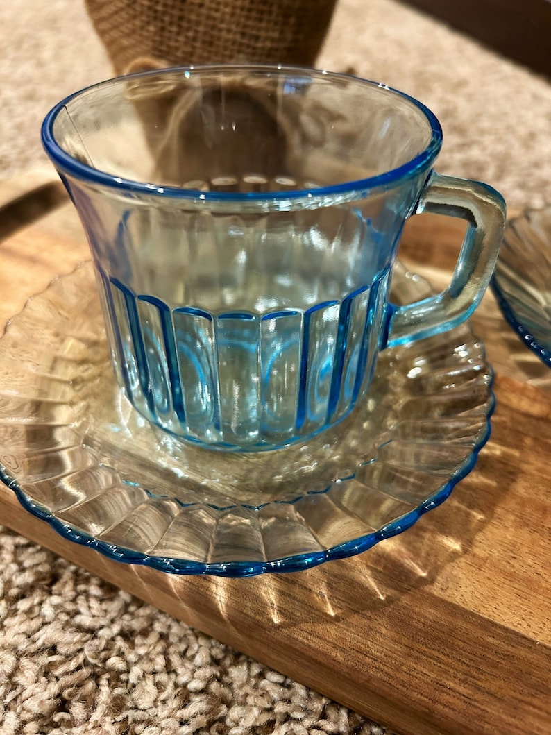 May include: A light blue glass teacup and saucer set. The cup has vertical lines and a handle. The saucer has a scalloped edge.