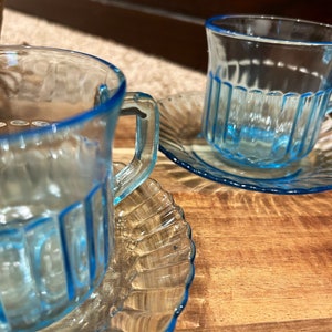 May include: Two blue glass teacups with matching saucers. The cups and saucers have a textured, ribbed design and a blue rim.