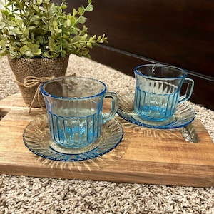 May include: Two light blue glass teacups and saucers with a ribbed design. The cups and saucers are on a wooden cutting board.