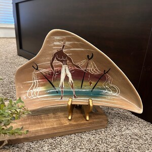 May include: A brown ceramic serving dish with a stylized image of a person holding a net. The dish is shaped like a crescent moon and has a white background with colorful accents. The dish is on a wooden stand.