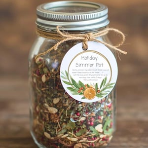 May include: A clear glass jar filled with dried herbs and spices, topped with a silver lid and tied with twine. A round tag reads "Holiday Simmer Pot" with instructions and a festive design including greenery and an orange slice.