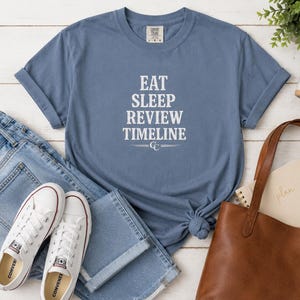 May include: A blue short-sleeve t-shirt featuring the white text EAT SLEEP REVIEW TIMELINE with a CC logo and horizontal lines underneath. The shirt is displayed alongside blue denim jeans, white canvas sneakers, and a brown leather tote bag.