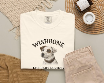 May include: A cream-colored short-sleeve t-shirt featuring a graphic of a Jack Russell Terrier dog. Above the dog is the text WISHBONE in a dark brown arch, and below the dog is the text LITERARY SOCIETY in dark brown capital letters.