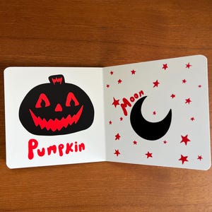 May include: A children's book open to two pages. One page features a black pumpkin with a red, jagged mouth and the word "Pumpkin" in red. The other page shows a black crescent moon and red stars with the word "Moon".