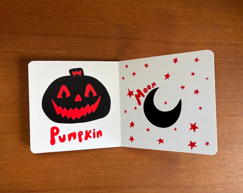 May include: A children's book open to two pages. One page features a black pumpkin with a red, jagged mouth and the word "Pumpkin" in red. The other page shows a black crescent moon and red stars with the word "Moon".