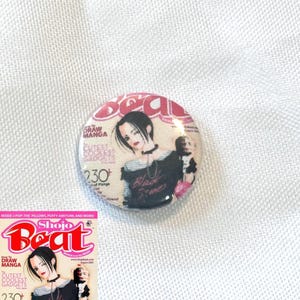 May include: A round button featuring a manga illustration with the text "Black Stones" and "Nana". The button has a pink and white background with the text "Shojo Beat" and "Draw Manga".