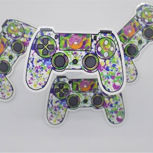 May include: A collection of colorful controller stickers. The stickers feature a white background with a splatter paint design in purple, green, blue, and yellow. Each controller has a donut graphic in the center.
