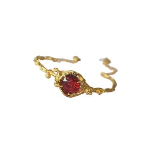 May include: A gold-toned bracelet featuring a central round, faceted deep red gemstone. The band has a textured, organic appearance resembling intertwined twigs or branches, with the stone held in place by small, delicate metal prongs.