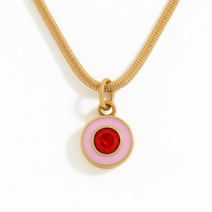 May include: A gold-toned snake chain necklace featuring a circular pendant. The pendant has a light pink enamel border surrounding a central, faceted, circular red gemstone. The entire piece is crafted from a metallic gold-coloured material.