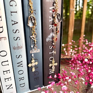 May include: Gold-toned metal and crystal bookmarks hanging from the top of a row of books. The bookmarks feature clear crystals, pearls, and ornate crosses. The books have titles like "Glass Sword" and "War Storm". Pink flowers are in the foreground.