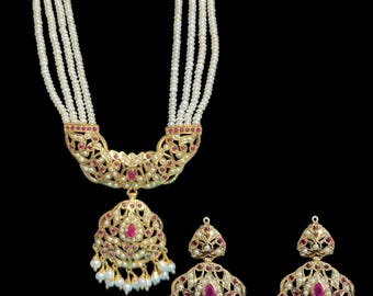 Op de afbeelding: Een gouden ketting met een hanger en bijpassende oorbellen. De ketting is gemaakt van meerdere strengen witte parels en heeft een gouden hanger met roze edelstenen. De oorbellen zijn ook van goud en hebben roze edelstenen.
