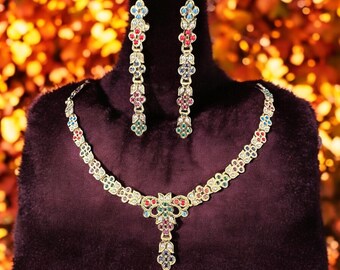 Op de afbeelding: Een gouden ketting en oorbellenset met een bloemmotief en gekleurde edelstenen. De ketting heeft een hanger met een cluster van groene, rode en blauwe stenen.