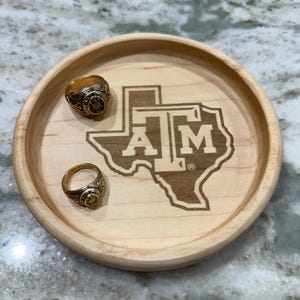 May include: A round, light-colored wooden tray with two gold rings and a laser-etched Texas A&M University logo. The tray is made of light-colored wood and has a simple, clean design. The rings are gold and feature intricate details.