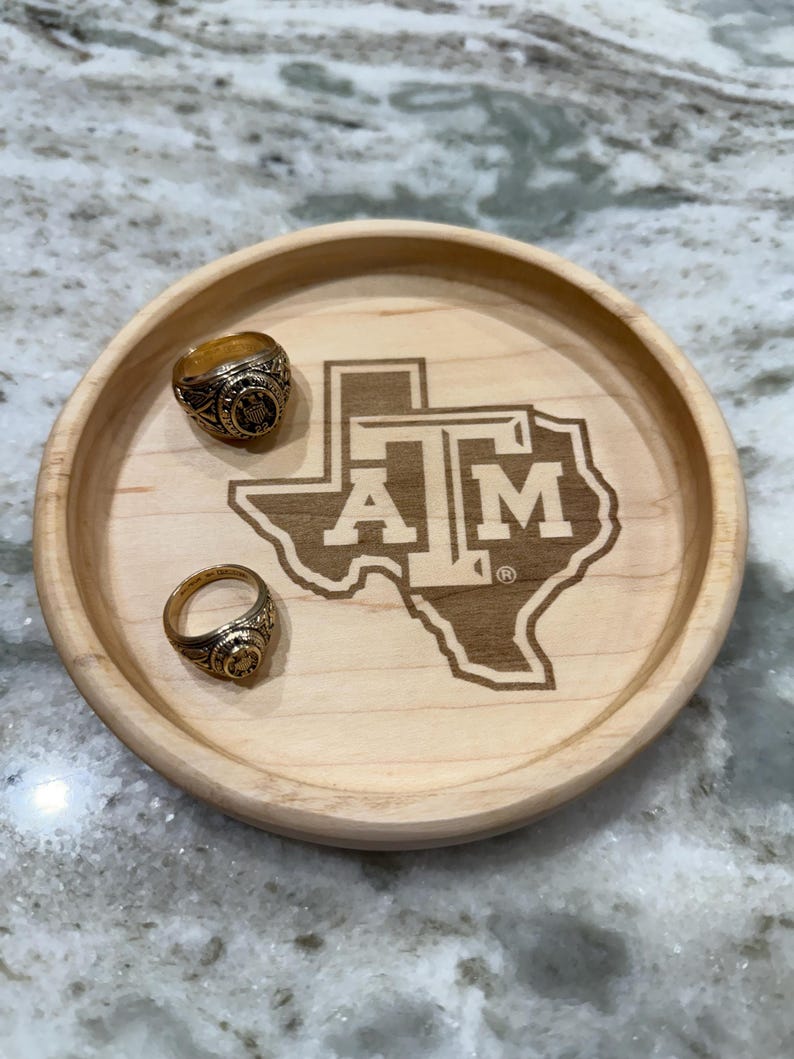 May include: A round, light-colored wooden tray with two gold rings and a laser-etched Texas A&M University logo. The tray is made of light-colored wood and has a simple, clean design. The rings are gold and feature intricate details.