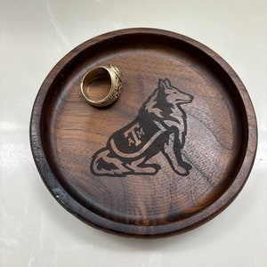 May include: A round, dark brown wooden tray with a laser-etched wolf design and the letters "A&M". A gold ring rests on the tray. The tray is likely used for holding small items.
