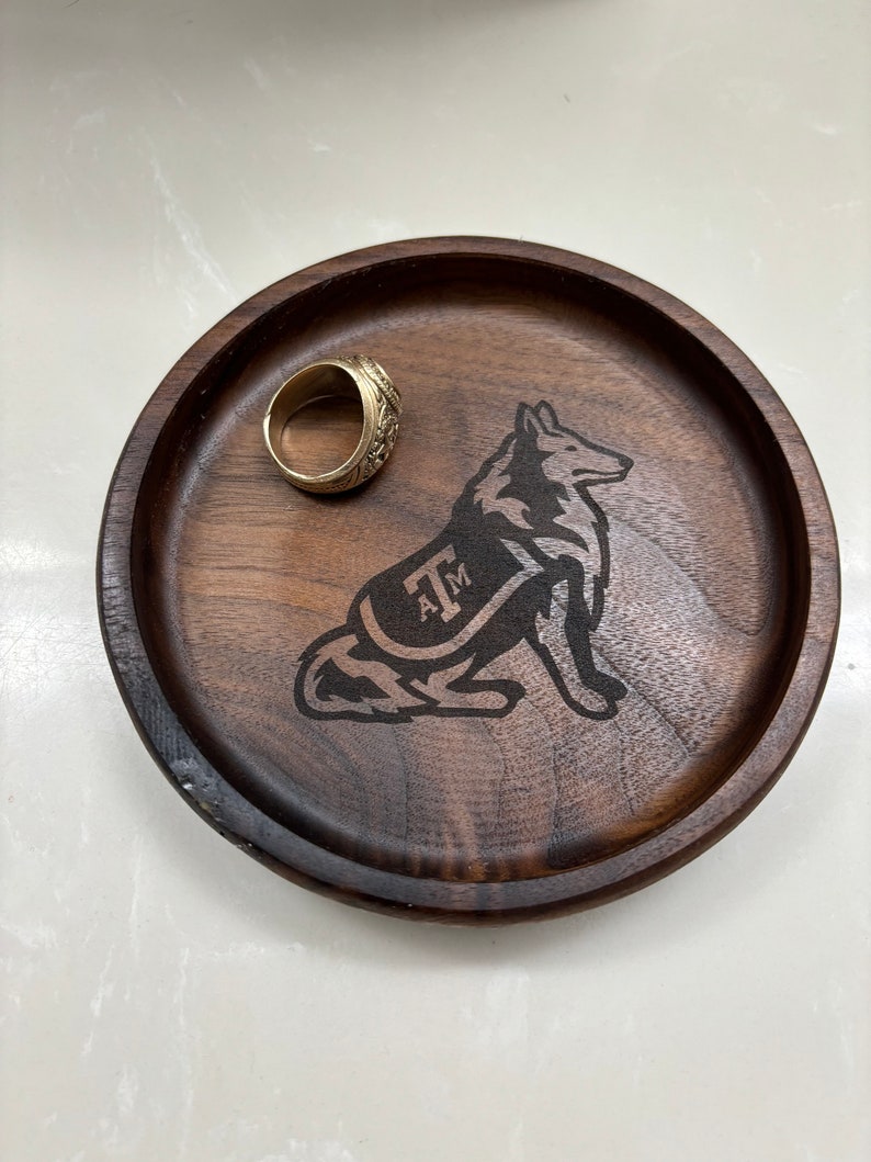 May include: A round, dark brown wooden tray with a laser-etched wolf design and the letters "A&M". A gold ring rests on the tray. The tray is likely used for holding small items.