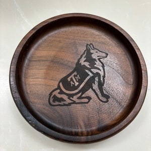 May include: A round, dark brown wooden tray with a carved image of a dog wearing a vest with the letters "A&M". The tray has a raised edge and a smooth surface. The dog is in a sitting position.