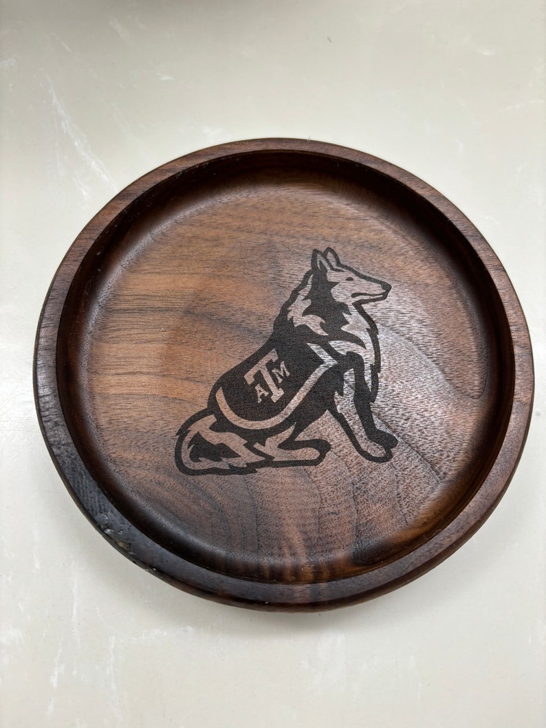May include: A round, dark brown wooden tray with a carved image of a dog wearing a vest with the letters "A&M". The tray has a raised edge and a smooth surface. The dog is in a sitting position.
