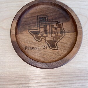 May include: A round, wooden tray with a carved Texas A&M University logo and the name "Frances '21". The tray is made of dark brown wood and has a raised edge.