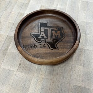 May include: A round, wooden coaster with a dark brown finish. The coaster features a laser-etched design of the Texas A&M logo within the outline of the state of Texas, along with the text "Caleb '24".