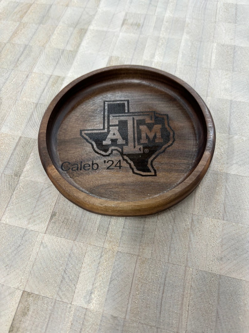 May include: A round, wooden coaster with a dark brown finish. The coaster features a laser-etched design of the Texas A&M logo within the outline of the state of Texas, along with the text "Caleb '24".