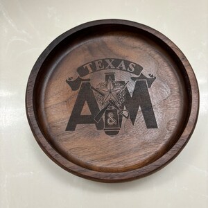 May include: A round, dark brown wooden tray with a raised edge. The tray features the engraved text "TEXAS A&M" with a star and other symbols. The wood grain is visible, adding texture to the design.
