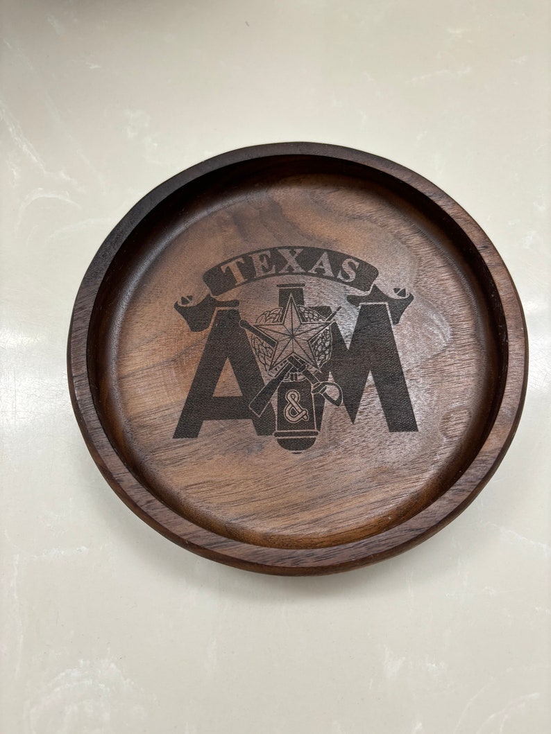 May include: A round, dark brown wooden tray with a raised edge. The tray features the engraved text "TEXAS A&M" with a star and other symbols. The wood grain is visible, adding texture to the design.