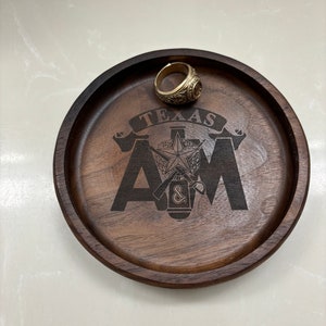 May include: A round, dark brown wooden tray with the text "TEXAS A&M" and a star emblem etched into the surface. A gold-colored ring with intricate detailing rests on the tray. The tray is on a white surface.
