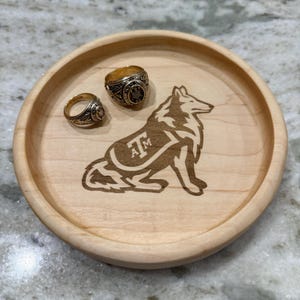 May include: A round, light-colored wooden tray with a carved wolf design and the letters "AM". Two gold rings rest on the tray. The tray is approximately 6 inches in diameter.