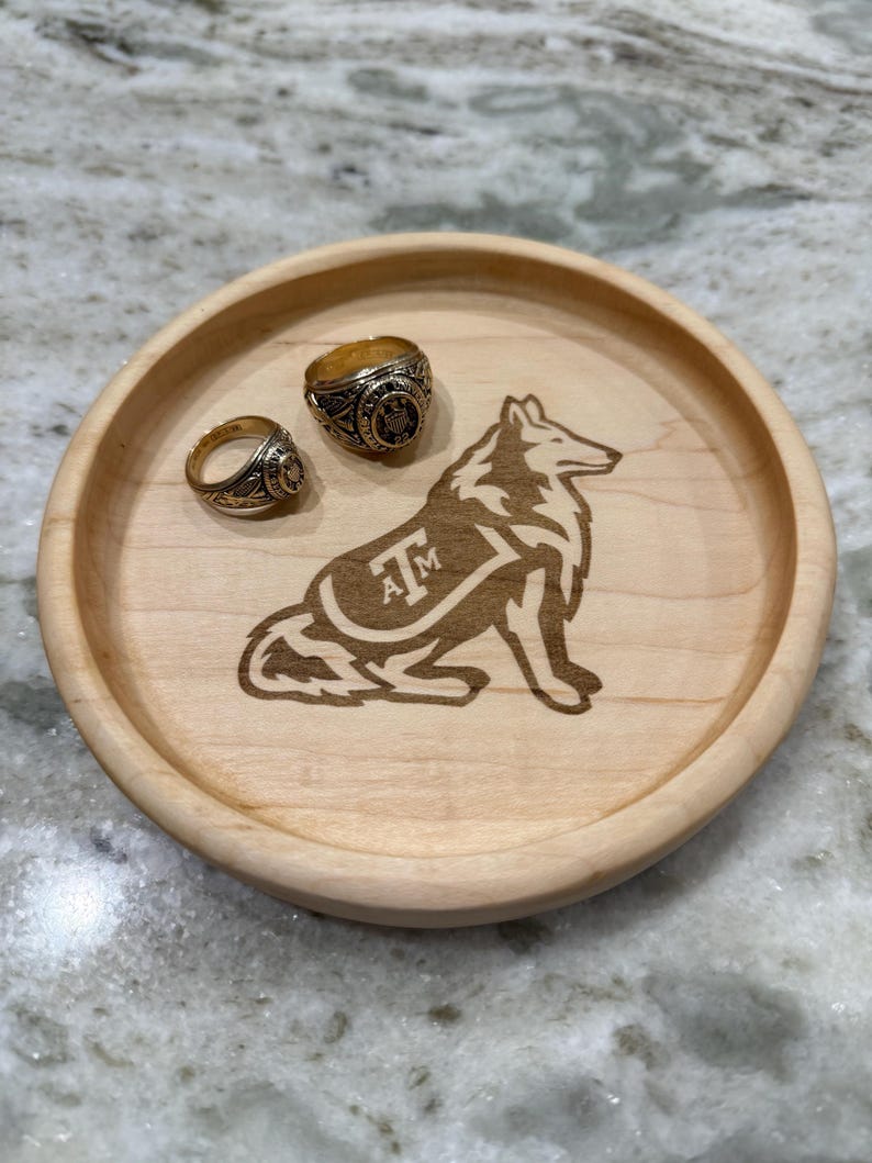 May include: A round, light-colored wooden tray with a carved wolf design and the letters "AM". Two gold rings rest on the tray. The tray is approximately 6 inches in diameter.