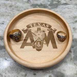 May include: A wooden tray with the engraved text "TEXAS A&M" and two gold rings. The tray is round and made of light-colored wood. The rings have intricate designs and are placed on the tray.