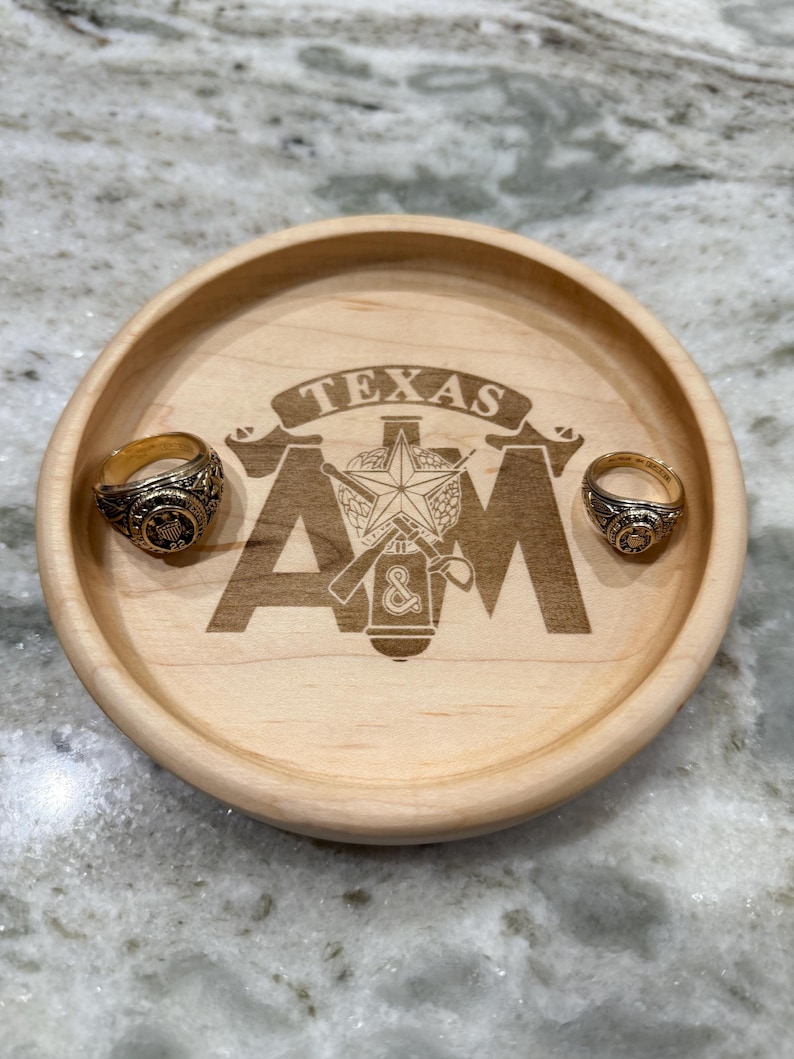 May include: A wooden tray with the engraved text "TEXAS A&M" and two gold rings. The tray is round and made of light-colored wood. The rings have intricate designs and are placed on the tray.