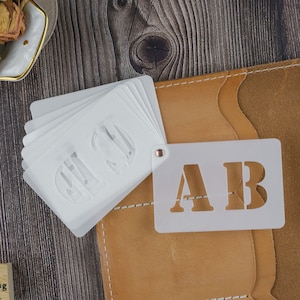 May include: A set of white plastic stencils with the letters "AB" and "HI" cut out. The stencils are on a brown leather surface with a white background.