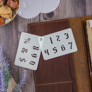May include: Two white plastic stencils with numbers and symbols. The stencils are on a brown leather journal with a pen and other craft supplies.