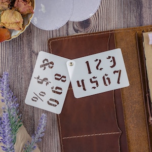 May include: Two white plastic stencils with numbers and symbols. The stencil on the left has the symbols %, $, and numbers 0-9. The stencil on the right has numbers 1-7. The stencils are on a brown leather journal with a pen and other craft supplies.