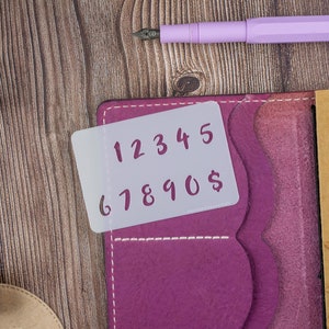 May include: A white plastic stencil with the numbers 1 through 9 and a dollar sign printed in pink. The stencil is placed on a pink leather journal with a brown leather cover next to it.