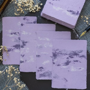 May include: Five sheets of lavender paper with a white and purple marbled design. The paper is stacked on a dark surface.