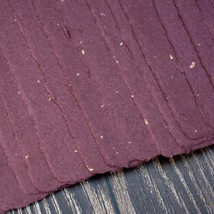May include: Close-up of handmade paper in a deep burgundy hue. The paper has a textured, layered appearance with visible fibers and flecks of gold. The edges are uneven, suggesting a handcrafted process. The paper is stacked diagonally against a dark wood surface.