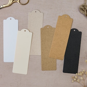 May include: A collection of blank rectangular bookmarks in various colors: white, cream, beige, brown, and black. Each bookmark has a rounded top with a small hole. Gold scissors and twine are also visible.