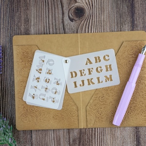 May include: Two white plastic stencils with the alphabet, A-M, on a brown folder with a paisley pattern. A pink pen is also on the folder.
