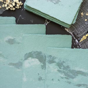 May include: A stack of light blue handmade paper sheets with a textured, marbled look. The paper is perfect for writing, crafting, or scrapbooking.