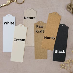 May include: A collection of rectangular paper tags in various colors: white, cream, natural, raw kraft, honey, and black. Each tag has a rounded top with a hole. Gold scissors, twine, and dried flowers are also present.