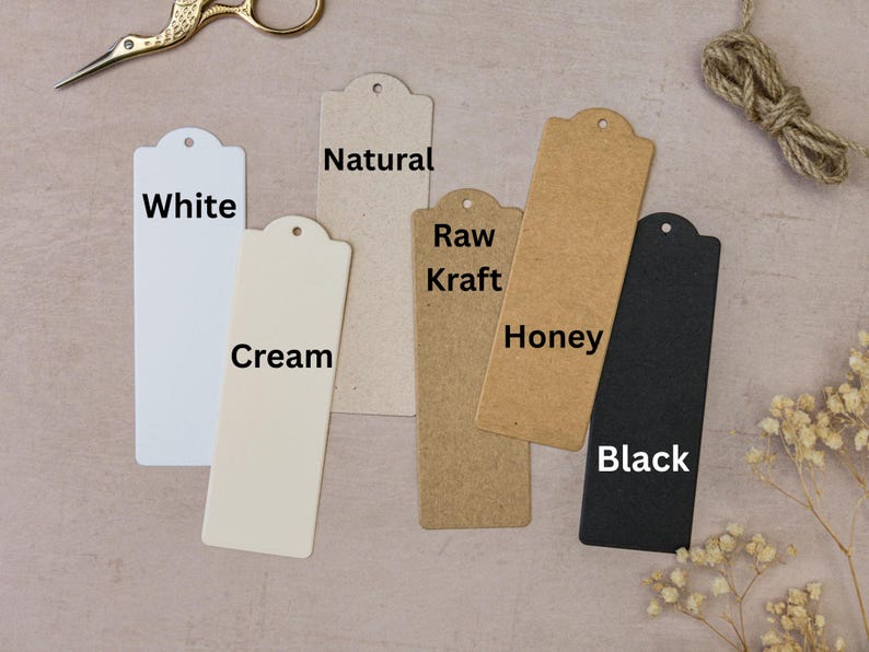 May include: A collection of rectangular paper tags in various colors: white, cream, natural, raw kraft, honey, and black. Each tag has a rounded top with a hole. Gold scissors, twine, and dried flowers are also present.