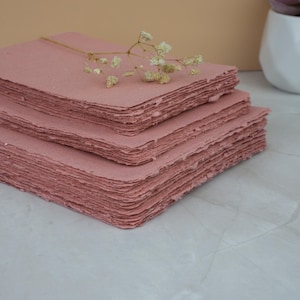 May include: Three stacks of handmade, rose-colored paper with rough, deckled edges. A sprig of delicate, white flowers rests on top. The paper is arranged on a light-colored surface, suggesting a craft or stationery theme.