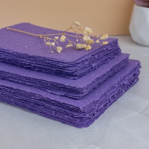 May include: A stack of handmade purple paper sheets with deckled edges. A sprig of dried white flowers rests on top. The paper has a textured appearance and is arranged in three layers.